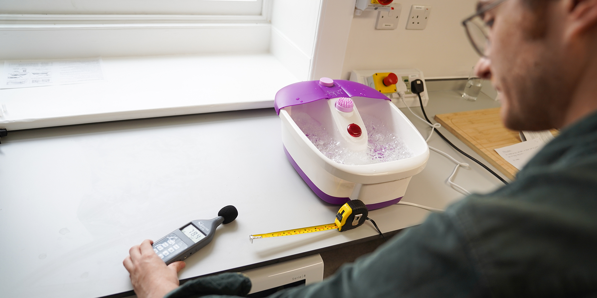Which? expert foot spa noise test: tester in green shirt placing decibel meter near foot spa, using tape measure to measure distance between the two