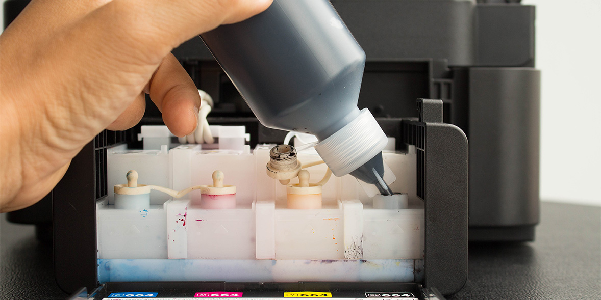 A hand holding a printer ink bottle, refilling the black ink tank of an Epson tank printer
