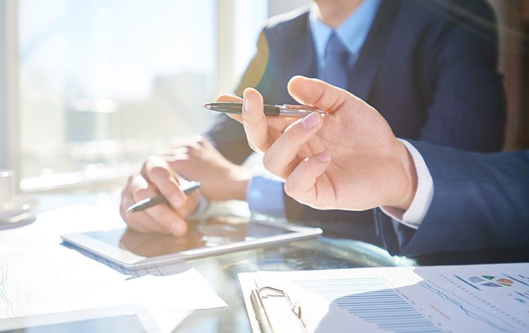 Two professionals in suits are engaged in a discussion at a table with documents and a tablet, one holding a pen.