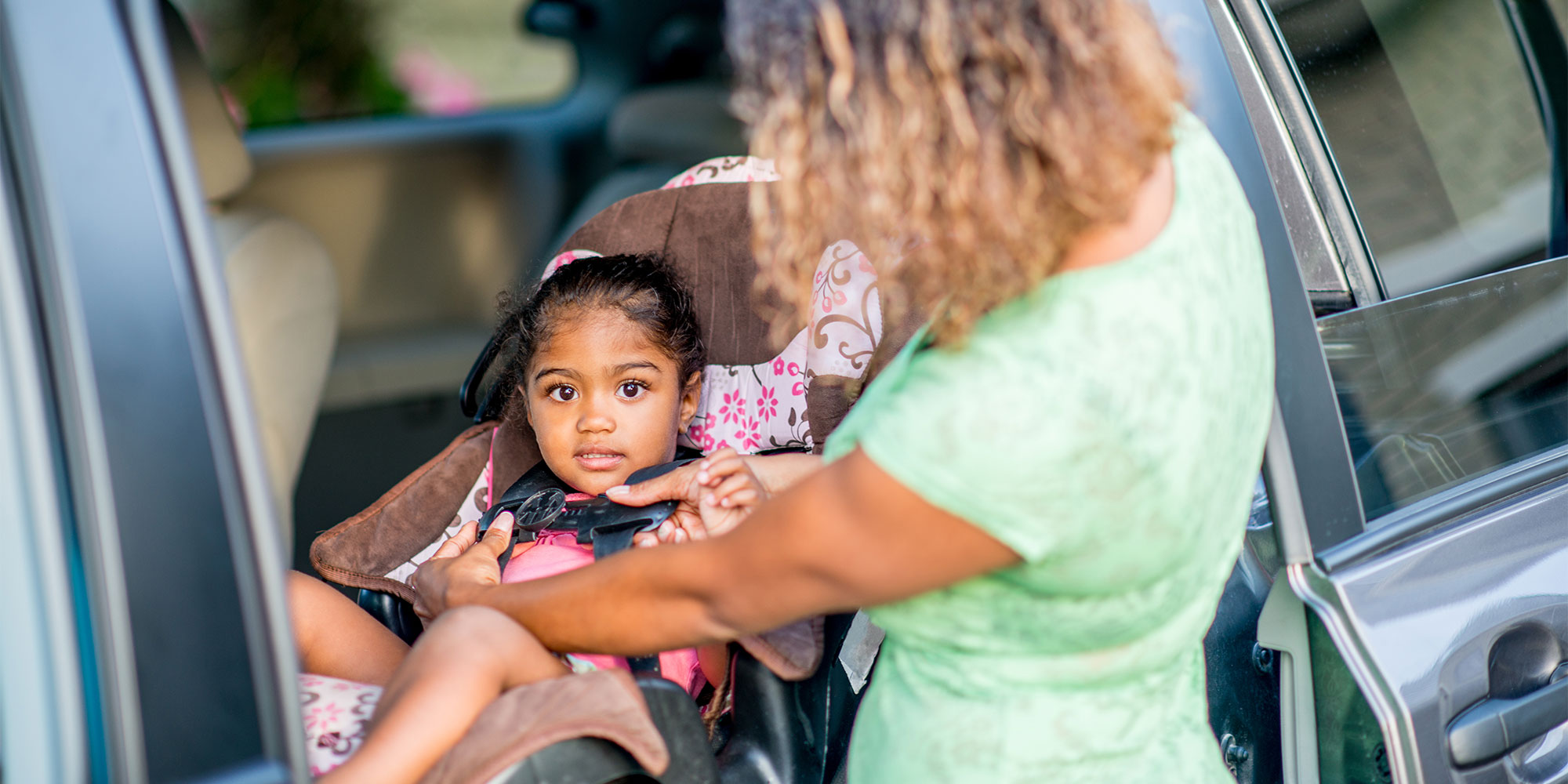 A woman stands outside an open car door, carefully adjusting the harness straps on a young girl sitting in a pink-patterned car seat.