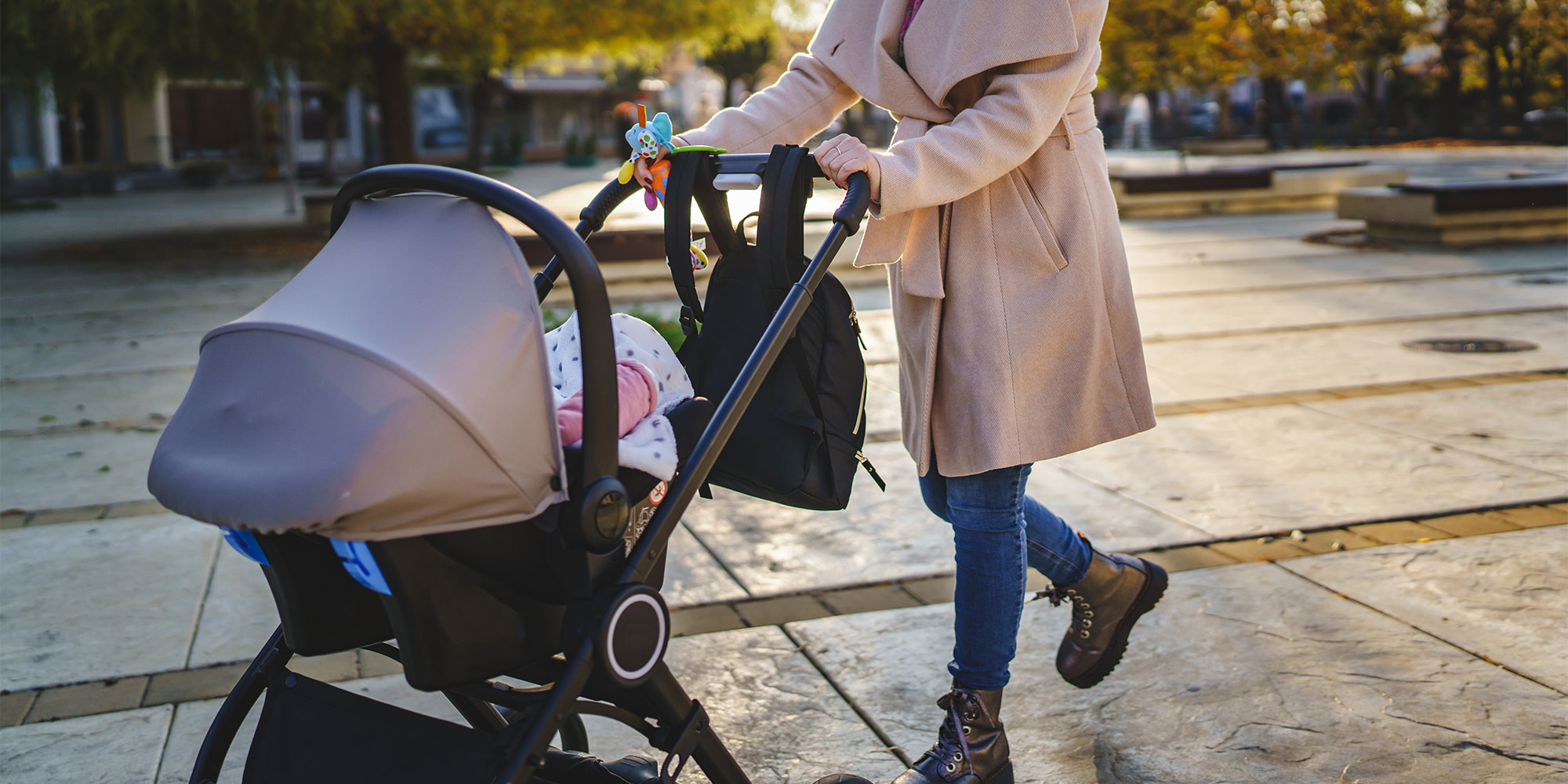 A person in a tan coat pushes a stroller across a stone-tiled plaza. A black backpack is hanging from the stroller handle, and a colorful toy is attached to the frame.