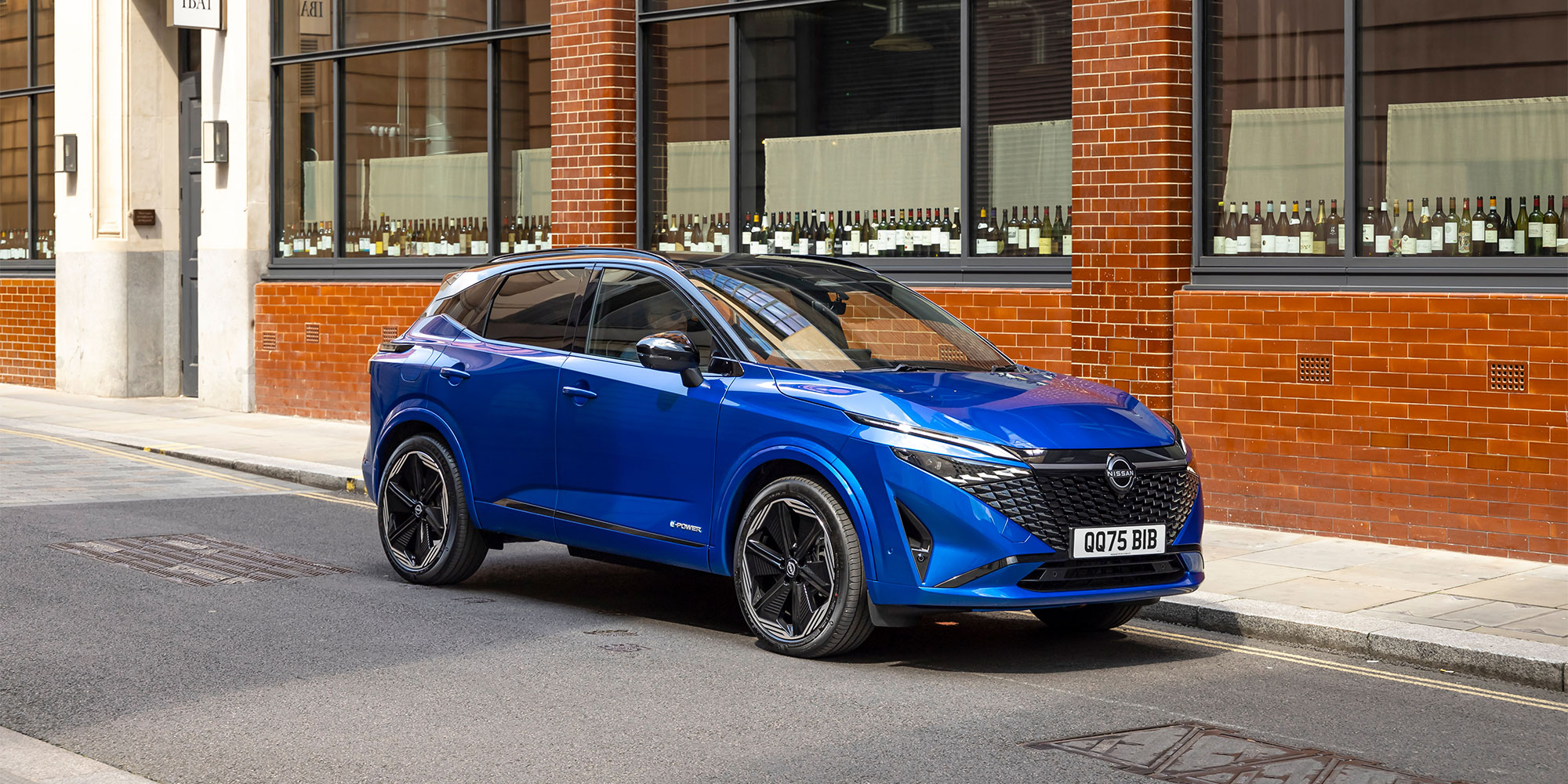 A blue Nissan Qashqai e-Power, parked on a street next to a building displaying several wine bottles.