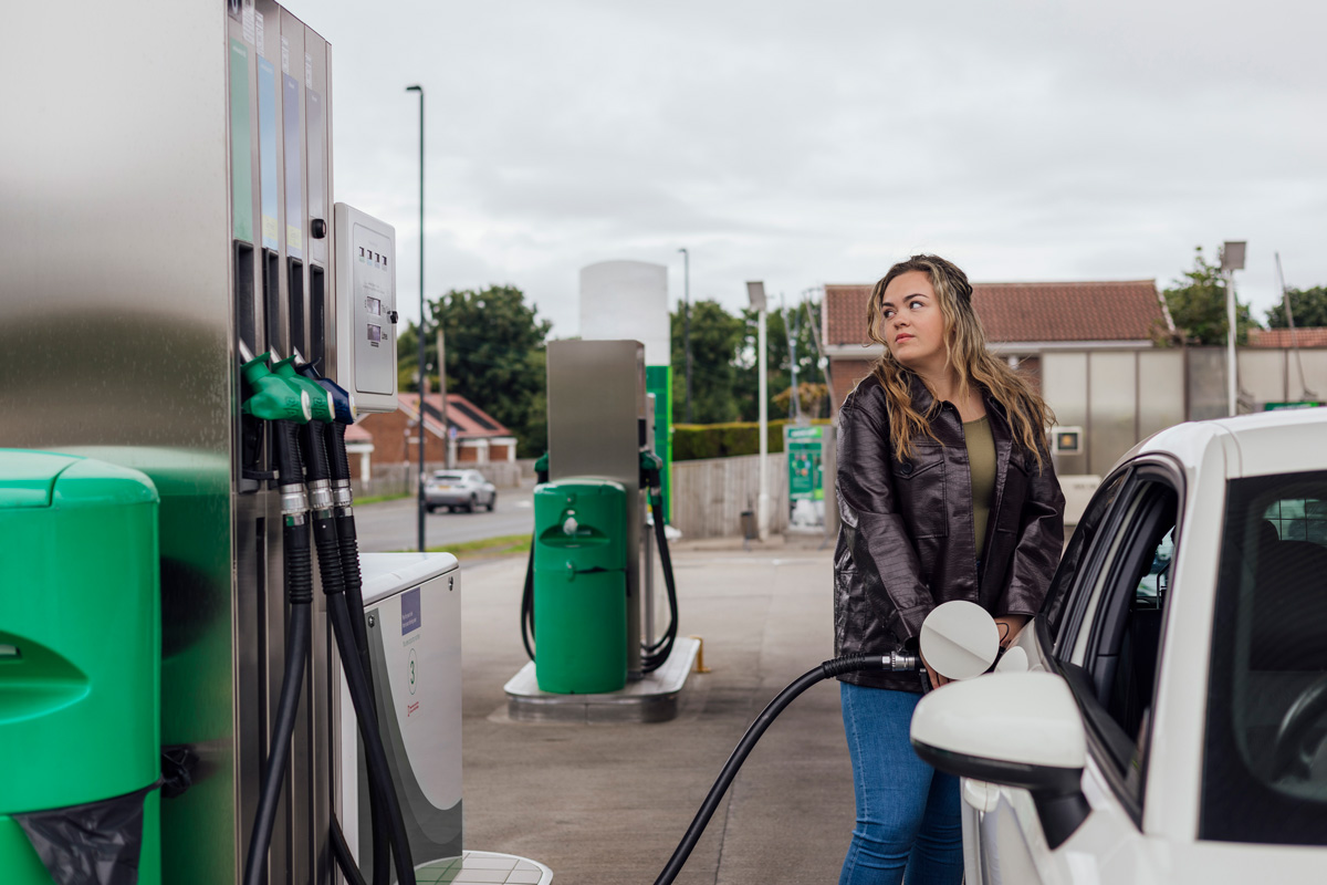 Woman in black jacket and blue jeans filling up white car at petrol station with green pumps.