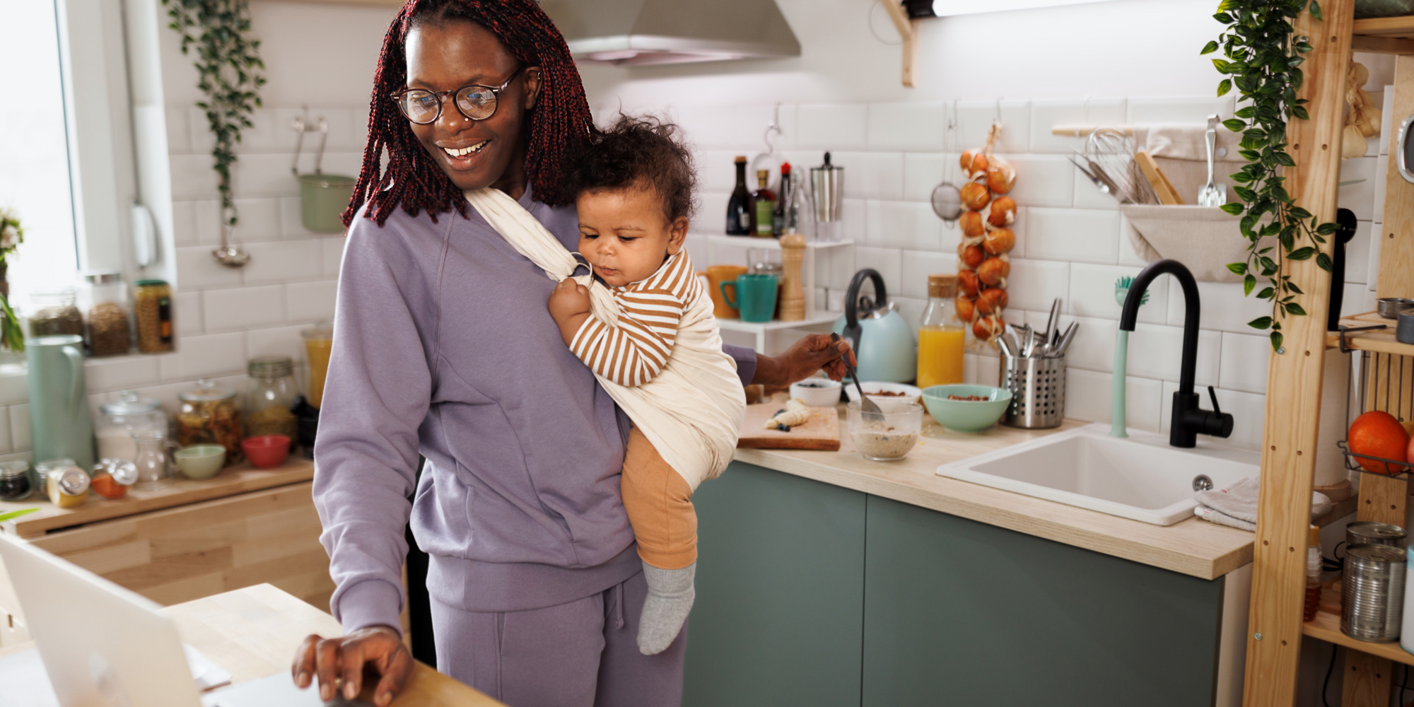 A woman wearing glasses and a purple tracksuit smiles as she uses a cream-coloured baby sling to carry a young child. She is in a kitchen, looking at a laptop while preparing food.