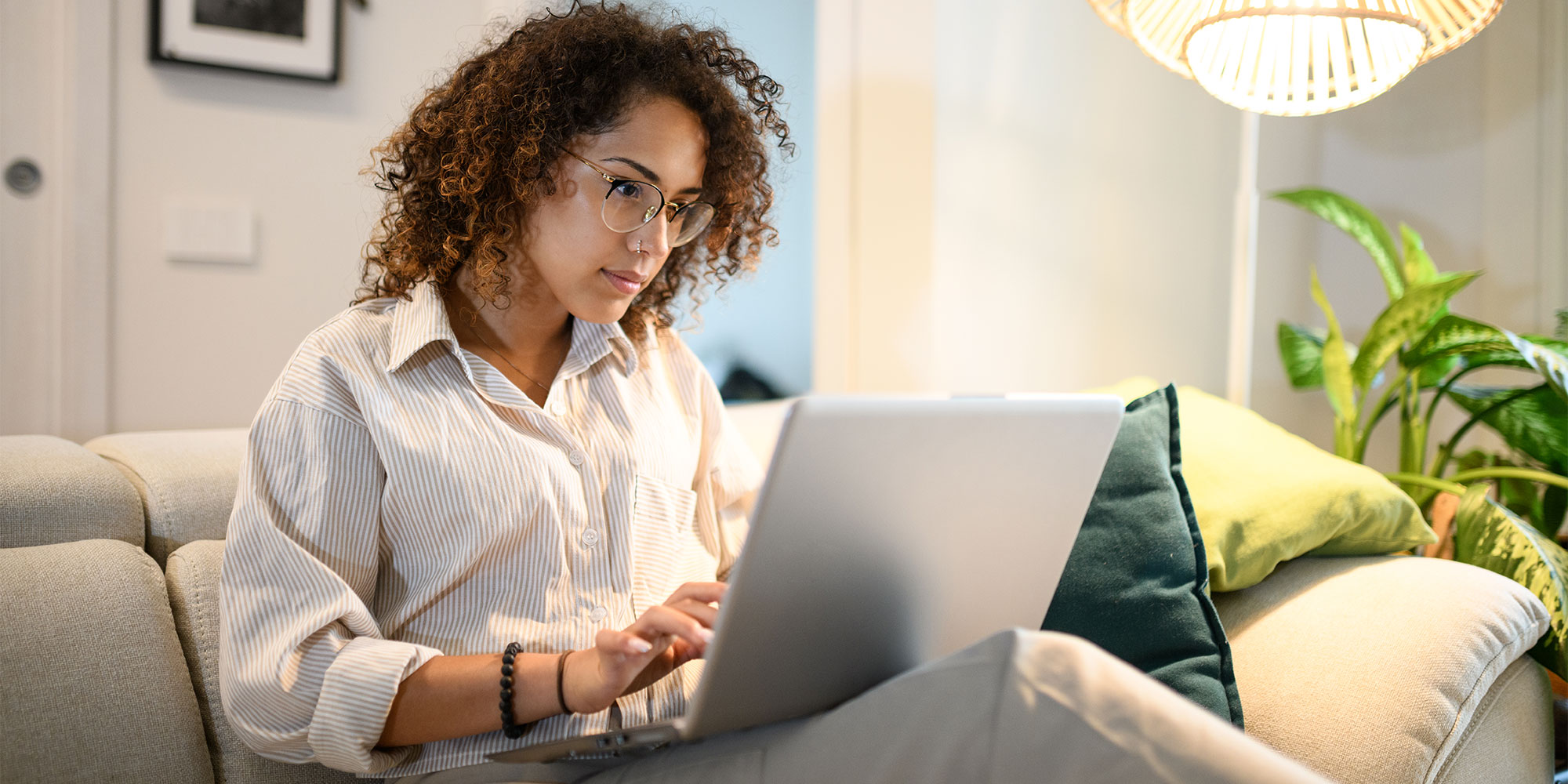Woman with curly hair wearing glasses and striped shirt using a laptop on a couch.
