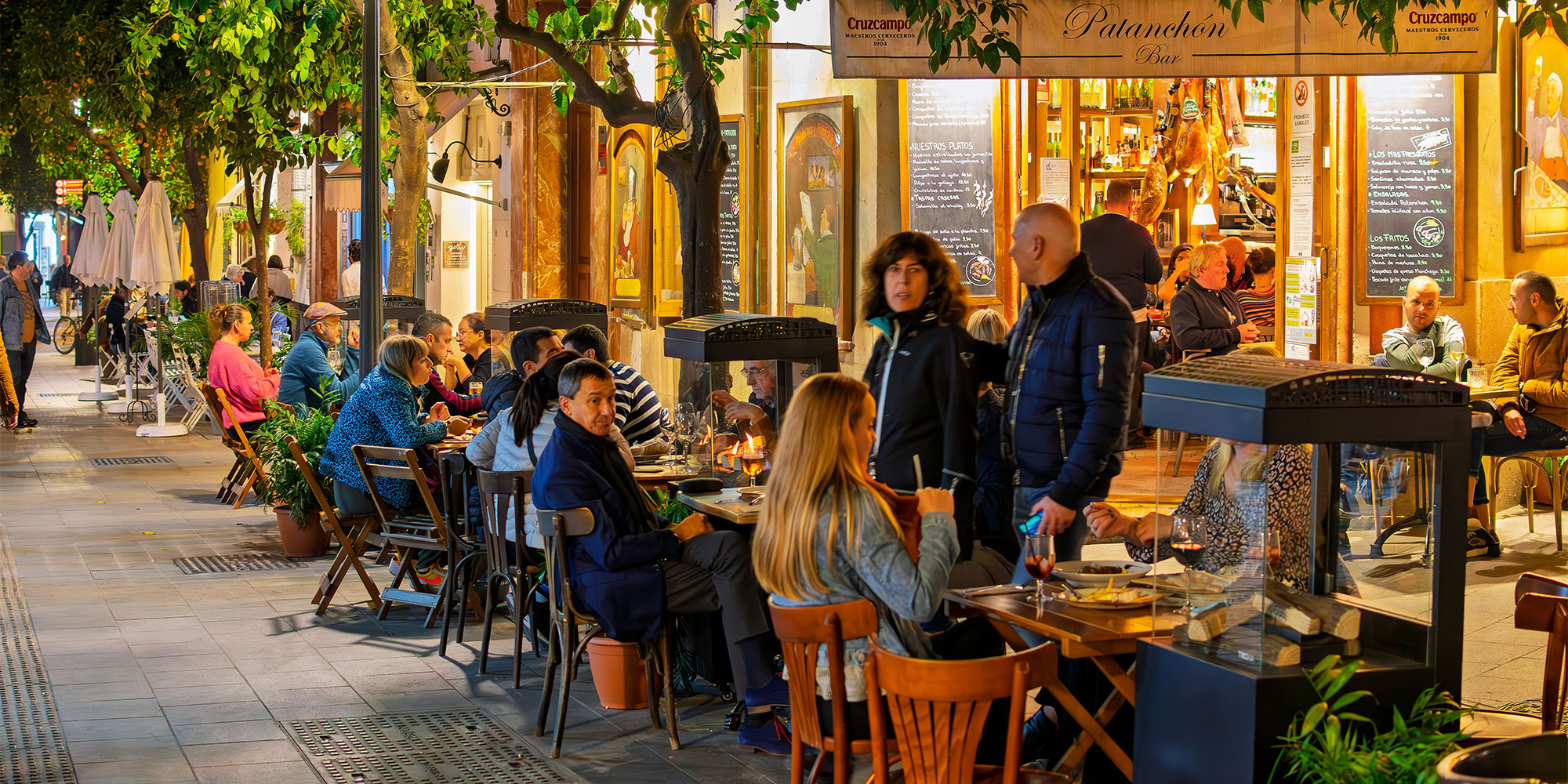 People dining outside a restaurant with outdoor heaters and potted plants on a sidewalk.