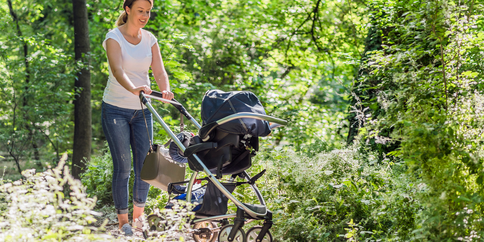 A woman in a white t-shirt and jeans pushes a stroller through a sunlit, wooded area with lush green foliage and wildflowers in the foreground.