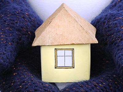 A small hand-painted house model with a brown roof, held between two hands wearing dark blue gloves.
